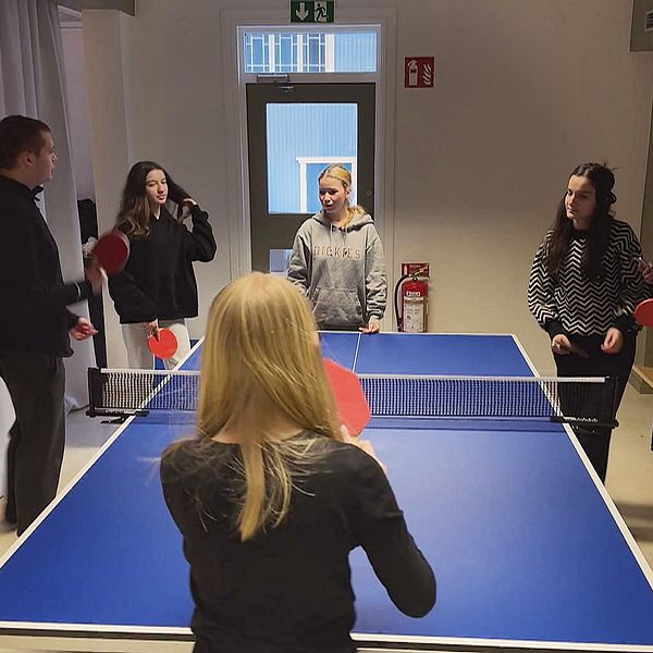 En grupp på fem personer spelar bordtennis i en inomhusmiljö. En person står på ena sidan av bordet och håller en racket, medan de andra fyra står på motsatt sida, beredda att spela. Rummen har ljusa väggar och en stor dörr i bakgrunden. Bänkar syns längs ena väggen.