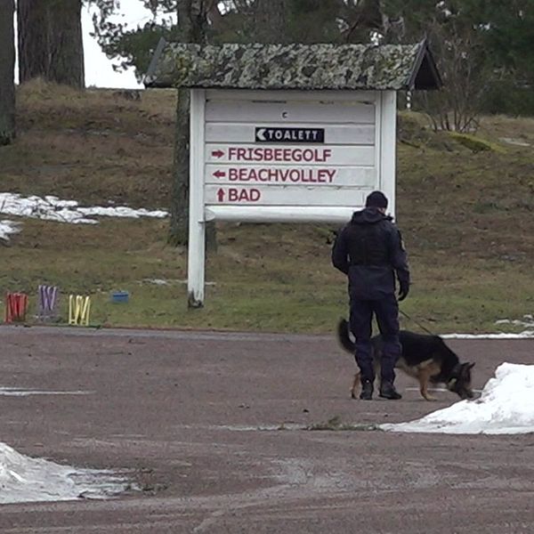 En polis går med en hund på en snötäckt väg bredvid en stor skylt som visar vägbeskrivningar till frisbeegolf, beachvolley och bad på en vintrig dag.