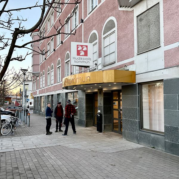 Entrén till Hudiksvalls kommuns byggnad med en skylt som visar kommunens namn. Två personer står vid ingången, med en cykel parkerad i bakgrunden. Fasaden är delvis i grått och rosa, och det är en klar himmel.