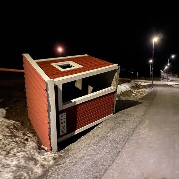 En välvt röd stuga som ligger på sidan vid en snötäckt väg, med gatubelysning som lyser upp omgivningen i mörkret.