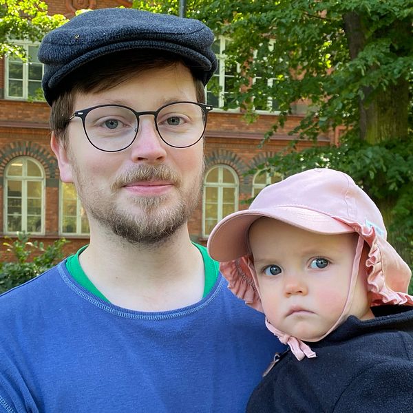 Man med keps och glasögon håller barn med rosa solhatt.