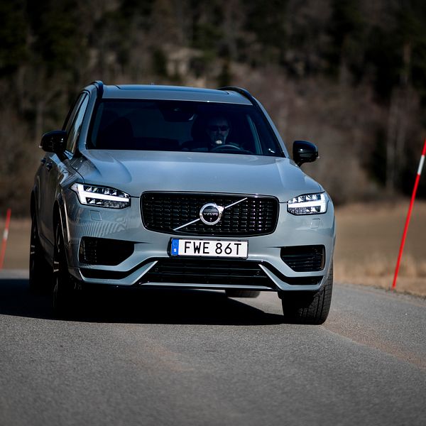 En Volvo XC90 körs på en landsväg med åkrar och skog i bakgrunden, med soligt väder vilket kastar skuggor på vägen. Registreringsskylten visar ”FWE 86T”.