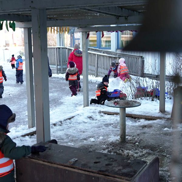 Barn i reflexvästar leker utomhus i en snöig förskolemiljö. Några barn springer och leker runt en lekställning, medan andra är sysselsatta vid en bänk. En del av bilden visar en struktur med en rund bordsskiva i förgrunden. Det är en kall och klar dag, och träden är utan löv.