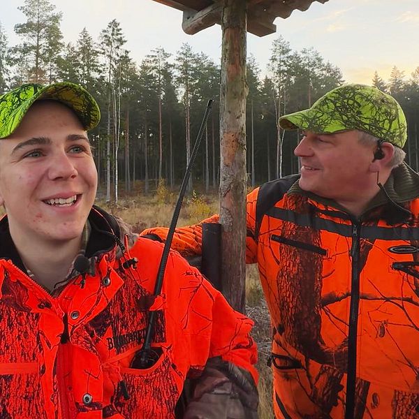 Två personer klädda i orange jaktkläder och matchande kepsar står i ett jakttorn med en skog i bakgrunden. Det är en solig dag, och omgivningen visar höga träd och en öppning i landskapet.