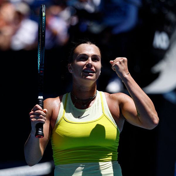 En tennisspelare i en gul tröja håller upp sitt racketslag, även medan hon visar entusiasm med en knuten näve. I bakgrunden syns åskådare som följer matchen.