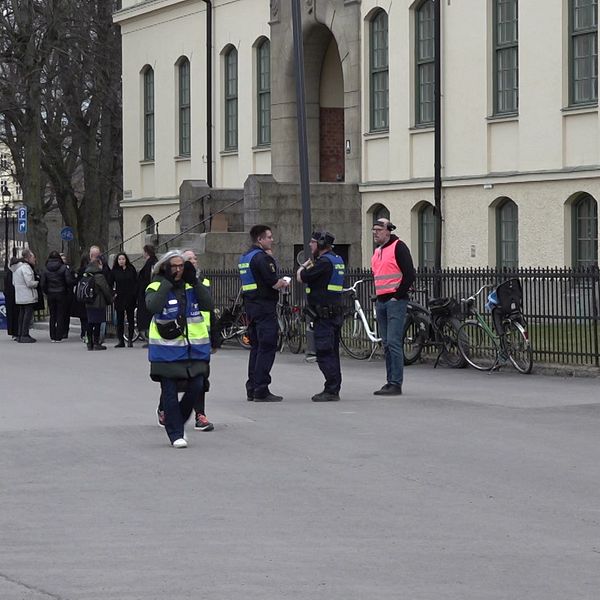 Säkerhetspersonal samlas nära en byggnad. En buss och några cyklar syns i bakgrunden.