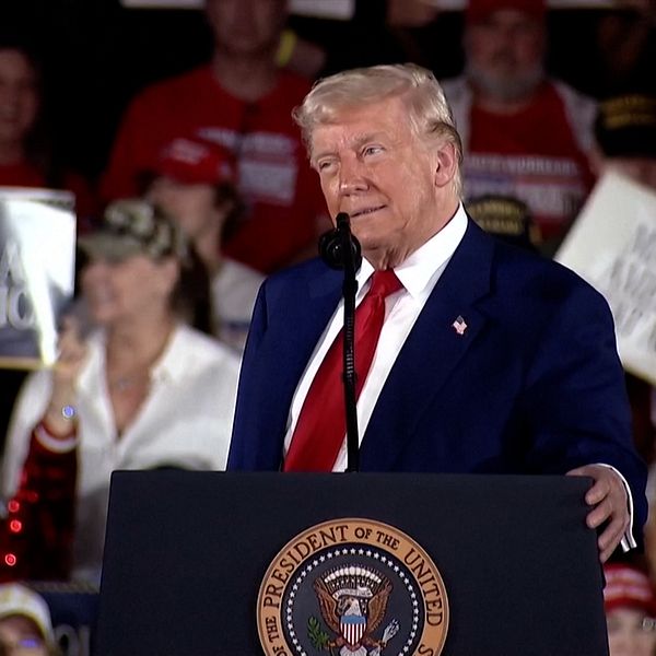 Politiker Donald Trump i kostym håller tal vid podium med presidentens sigill, publik i bakgrunden.