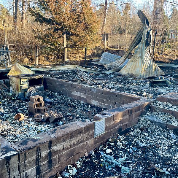 Resterna av en byggnad efter en brand, med nedbrunna väggar och aska spridd över området. Det syns en del av en takstruktur som har kollapsat, medan betongblock och brända föremål ligger kvar på marken. Området runtomkring är skogigt med några träd synliga.