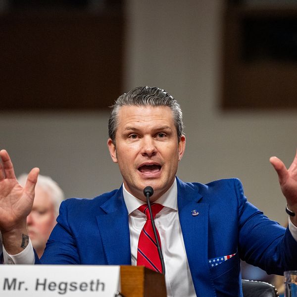 Pete Hegseth i en blå kostym med röd slips, gestikulerar med båda händerna. Främre bordet visar hans namn och titel ”Mr. Hegseth”. I bakgrunden syns suddiga figurer av andra åhörare.