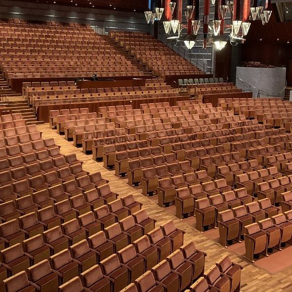 En inomhusarena med rader av tomma, bruna sitsar i ett modernt auditorium. Belysning och dekorativa detaljer syns i taket, vilket skapar en elegant atmosfär. Scenen är delvis synlig i bakgrunden, vilket indikerar att lokalen är avsedd för föreställningar eller evenemang.