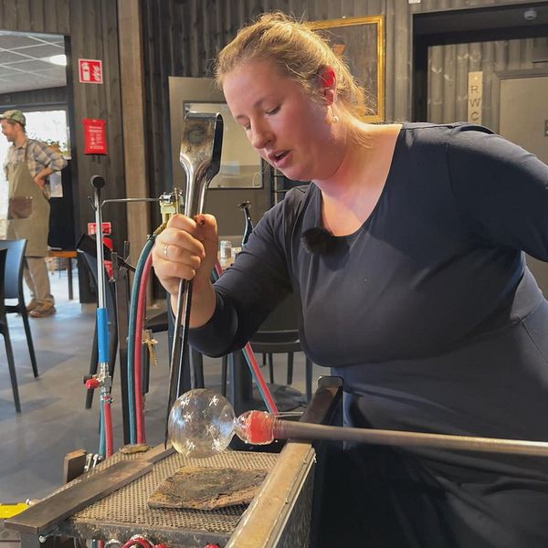 En person som arbetar med glasblåsning står vid en glasblåsarbänk. I förgrunden håller hon i ett glasverktyg och formar en glasboll. I bakgrunden syns två personer som tittar på, i en miljö med mörka väggar och moderna möbler.