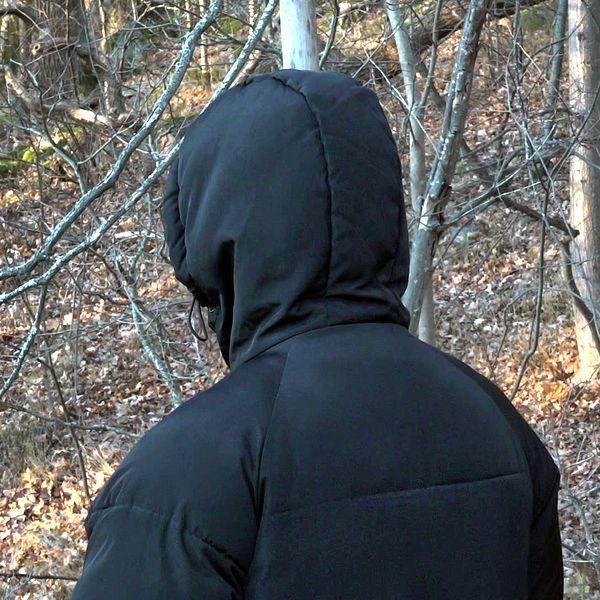 En person med svart jacka och huva står med ryggen vänd mot kameran, omgiven av skog med löv och träd.