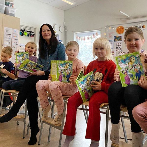 Barn sitter i en rad av stolar i en klassrumsmiljö, alla håller i böcker och ser engagerade ut. I mitten av raden sitter en vuxen. Rummet är dekorerat med färgglada affischer och material som tyder på en lärande miljö.