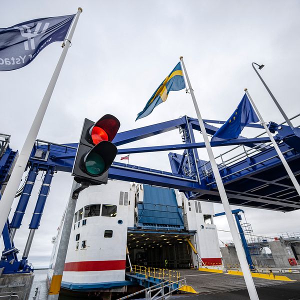 Färja som står vid en hamnterminal med nedfälld landgång för ombordstigning eller avstigning, omgiven av flaggor och ett trafikljus som visar rött ljus.