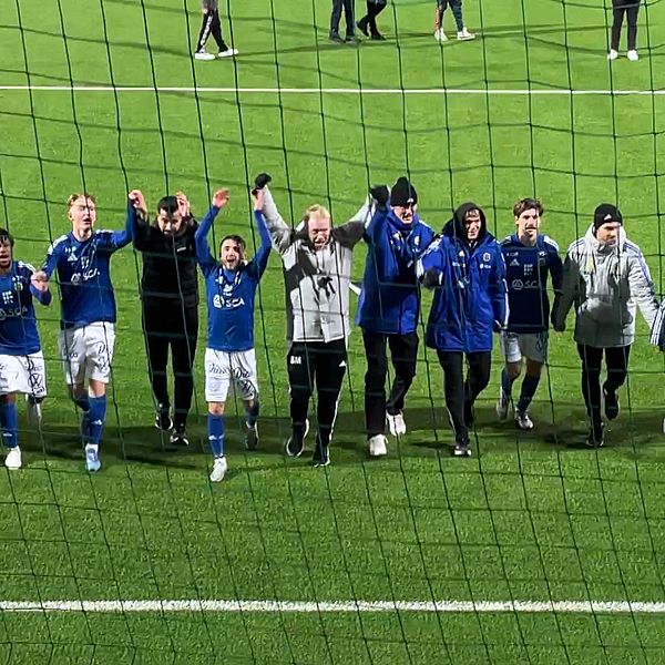 Lagmedlemmar och tränare firar en seger på en fotbollsplan. De står i rad framför en nätad målbur, klädda i blåa och svarta träningskläder, med händerna lyfta i luften och leenden på läpparna. I bakgrunden syns andra personer som deltar i firandet. Gräset är grönt och planen ser välskött ut.
