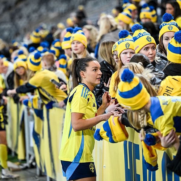 En samling av fans i gula och blå kläder, med matchande mössor, samlas vid en avspärrning för att heja och interagera med spelare efter en fotbollsmatch. Runt om dem finns ett festligt och energiskt stämning.