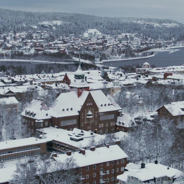 En panoramabild över en snötäckt stad med traditionell arkitektur, belägen vid en sjö. I förgrunden syns flera byggnader med snöbelagda tak, inklusive en stor, historisk byggnad med en tornspira. I bakgrunden reser sig en kuperad, snötäckt landskap med fler hus och träd, och sjön breder ut sig till höger.
