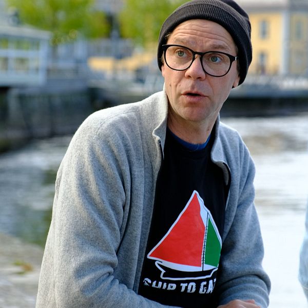 Person med grå kofta och svart t-shirt sitter utomhus vid vattnet, samtalar med annan person.