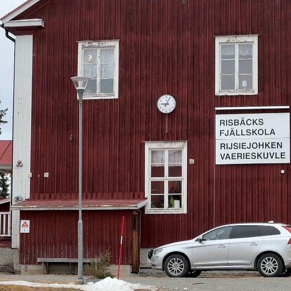 Röd byggnad, Risbäck fjällskola. Silverfärgad bil parkerad. Träd i bakgrunden.