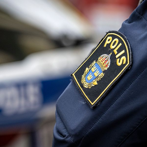 En närbild av en polisuniform där ett tydligt insynskt patch med texten ”POLIS” är synligt på ärmen. I bakgrunden syns en polisbil.
