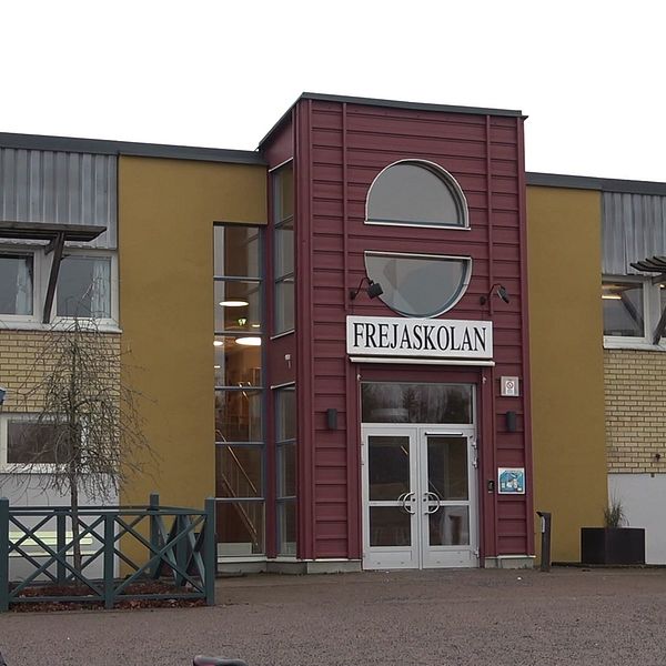 Fasaden av Frejaskolan, en modern byggnad med en röd och beige exteriör. Ingången är centralt placerad och omgiven av stora fönster. Omgivningen är lugn, med en grönskande gård och en trägrind synlig till vänster.