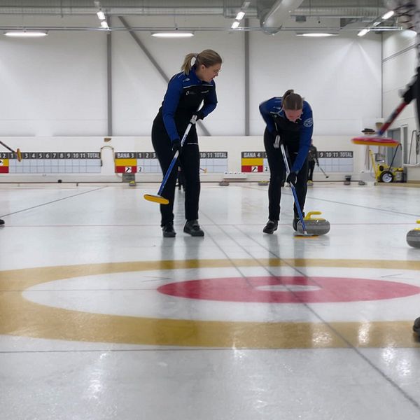Personer spelar curling på en inomhusbana. Sopare vid sidan om stenen.