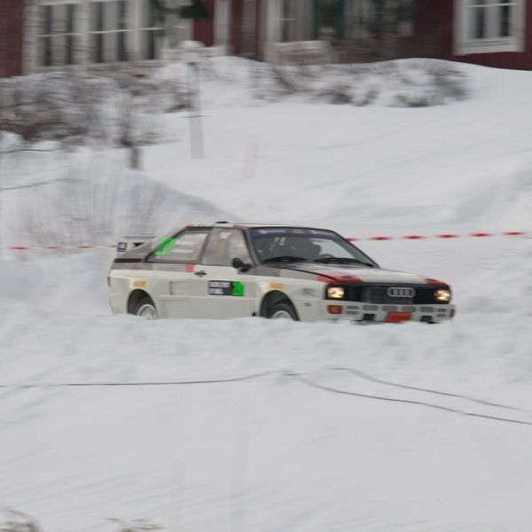 En rallybil kör genom ett snötäckt landskap i en vinterrallytävling. I bakgrunden syns ett par hus och träd täckta av snö.