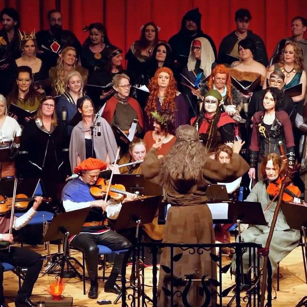Kör och orkester framför musik tillsammans på scen, många bär fantasifulla kostymer.
