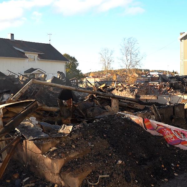 Detaljerad vy av en plats som drabbats av en brand, med jämna ruiner och nedbrunna byggnadsdelar. I bakgrunden syns en intakt byggnad och ett ljust blått himmel. Rester av bråte och aska är spridda över området, vilket vittnar om den förödelse som orsakats. En del av en färgglad skylt kan skymtas bland rubbningen.