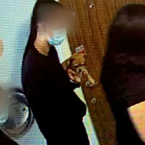 Tre personer, med ansiktena suddiga för anonymitet, står i en rad i en korridor utanför en dörr. De bär ansiktsmasker och kläder i mörka nyanser. Personen längst fram håller en mobiltelefon. En stor bokstav ”B” syns på väggen.