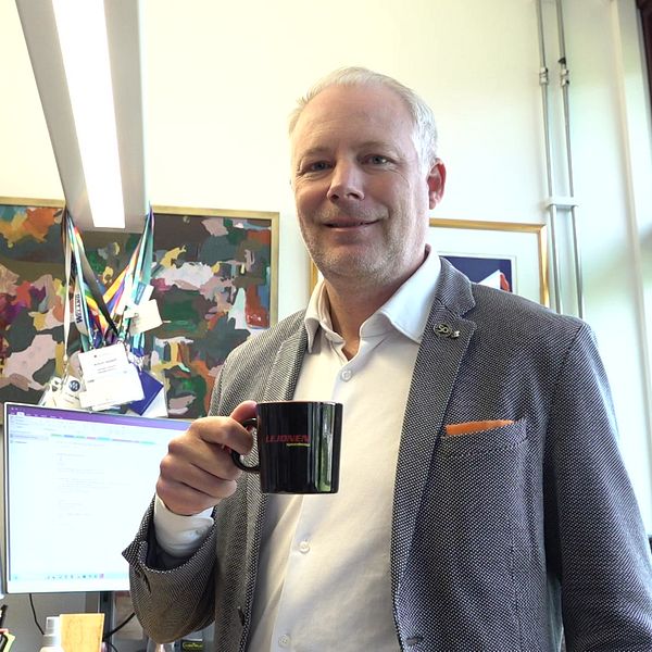 Politiker står vid ett skrivbord och håller en svart kopp med Lejonens logga på. I bakgrunden syns två datorskärmar som visar arbetsrelaterad information, tillsammans med konstverk på väggen. Rummet har stora fönster som släpper in naturligt ljus.