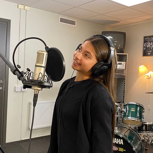 En person står framför en mikrofon i en inspelningsstudio, iklädd hörlurar. I bakgrunden syns ett trumset samt affischer på väggarna. Rummet har en professionell atmosfär med dämpad belysning.