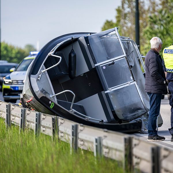 Bil har vält vid vägkant, polis och civilperson står bredvid, kö med bilar i bakgrunden.