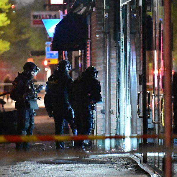 Tre poliser i mörka kläder står och ser på en skadad glasdörr.
