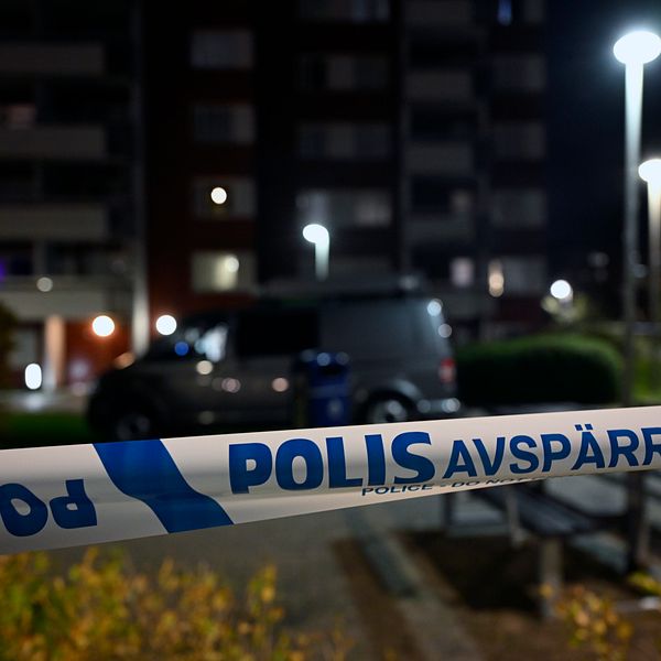 Polisavspärrning med texten ”POLIS AVSPÄRRAT” i förgrunden, med en bil och flerfamiljshus i bakgrunden, belyst av gatulampor under natten.