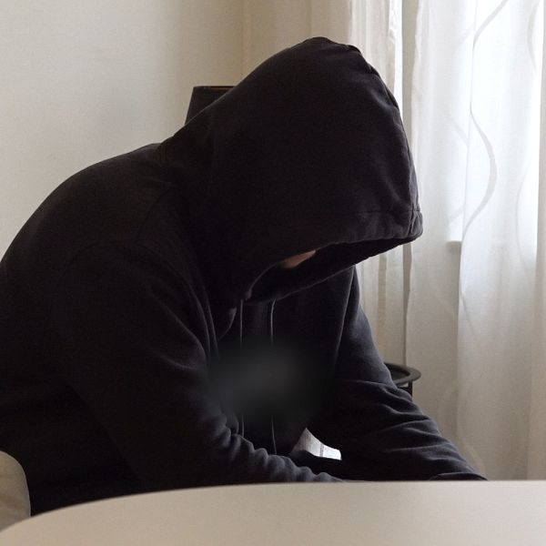 En person med huvtröja sitter på en soffa, lutad framåt med huvudet sänkt. Rummet har vitt ljus som strömmar in genom ett fönster, och en ljusstake syns på bordet framför. Det finns också en färgad kudde på soffan. Atmosfären verkar lugn och stillsam.