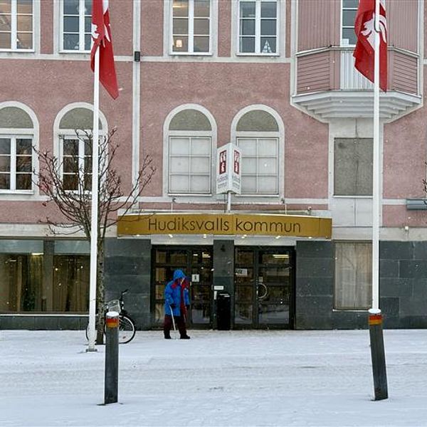 Entrén till Hudiksvalls kommunhus, där snö ligger på marken och två personer syns utanför. Klassiska fönster i grått och rosa syns på byggnadens fasad, med svenska flaggor som svajar.