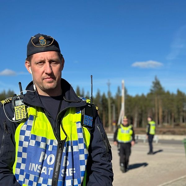 Polis med insatsväst står utomhus vid ett skogsområde, andra poliser i bakgrunden.