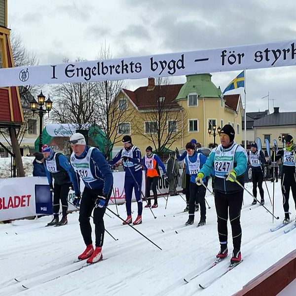 Skidåkare deltar i ett långlopp i en svensk stad, med en banderoll ovanför spåret. Flera åkare i nummerlappar syns i förgrunden, medan åskådare och färgglada byggnader finns i bakgrunden. En svensk flagga vajar bland träden.