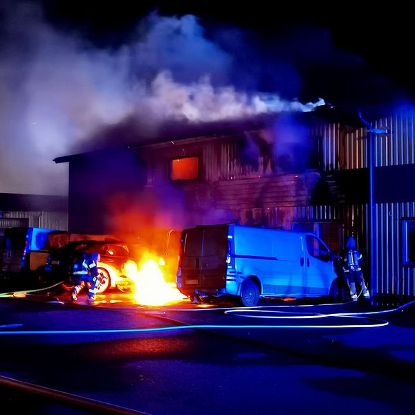 På bilden syns en brinnande bil. Det är mörkt ute. Bilen står utanför en industrilokal. En brandman är framme vid elden och försöker släcka den.