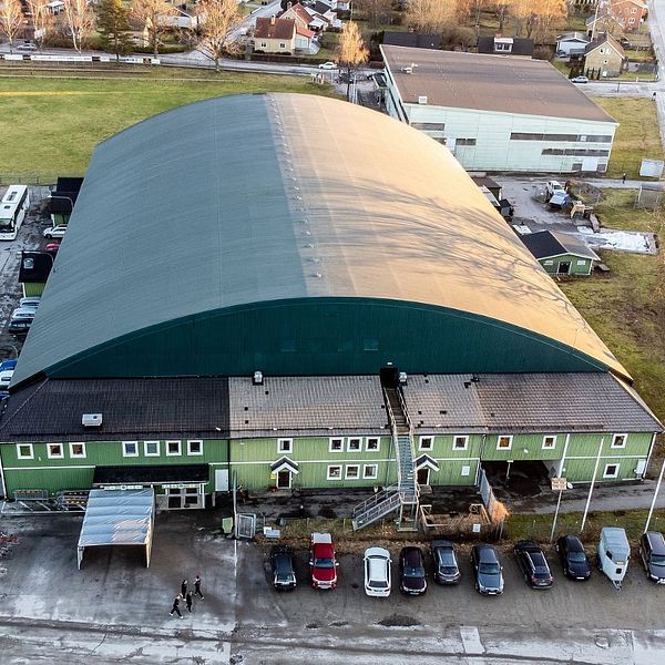 Grön sporthall med välvt tak, sett uppifrån. Flera bilar parkerade framför entrén.