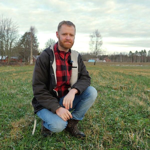 En person sitter på knä i ett öppet fält med grön gräs och några spridda träd i bakgrunden. Himlen är delvis molnig och ger en stillsam atmosfär. I bakgrunden syns några hus på avstånd.