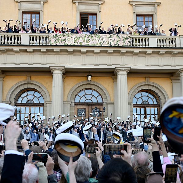 Studenter firar utanför skolbyggnad, viftar med mössor mot balkong.