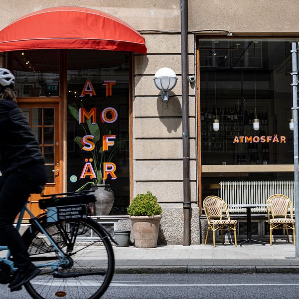 En cyklist passerar en restaurang med namnet ”ATMOSFÄR” som har en röd markis och utomhusmöbler. Fönstren har dekorativa ljus och en grön växt är placerad vid entrén.