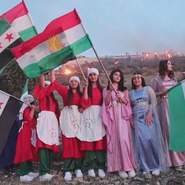 Grupp personer i festkläder håller kurdiska och syriska flaggan.