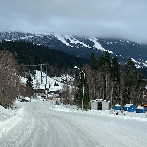 Snöig landsväg leder mot skidbackar och berg täckta av snö under molnig himmel.