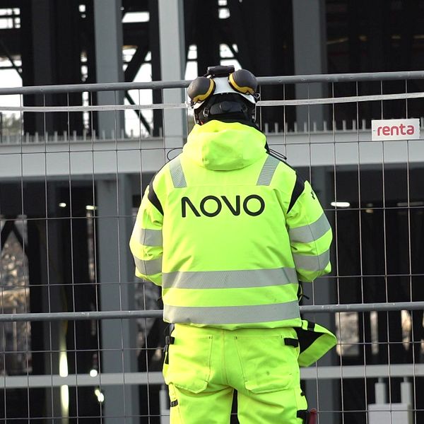 Byggarbetare i reflexväst står vid en avspärrning framför en byggnad under konstruktion vid bygget av Volvo Cars satsning på batterifabrik i Göteborg.
