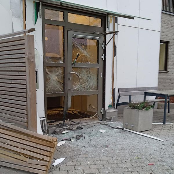 Skadad entré med spruckna fönster och en trasig dörr, där en del av fasaden har lossnat. Det ligger glas och skräp på marken, och en liggande träpanel är synlig vid sidan av dörren. I bakgrunden skymtar en grön växt i en kruka och byggnadens väggar.