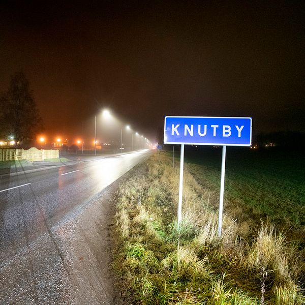 Skylt vid vägkanten med texten Knutby i mörker och billyktor i bakgrunden