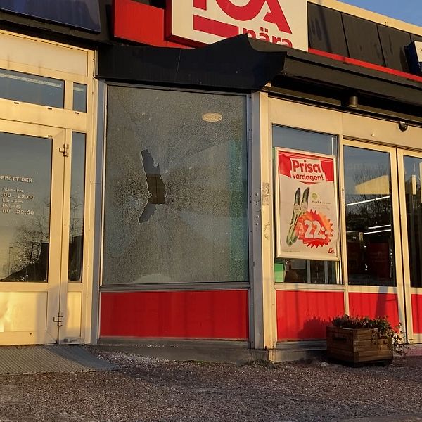 Krossad fönsterruta vid ingången till en Ica-butik med synliga skyltar och reklam. Butikens dörr är öppen och det finns snö på marken framför.